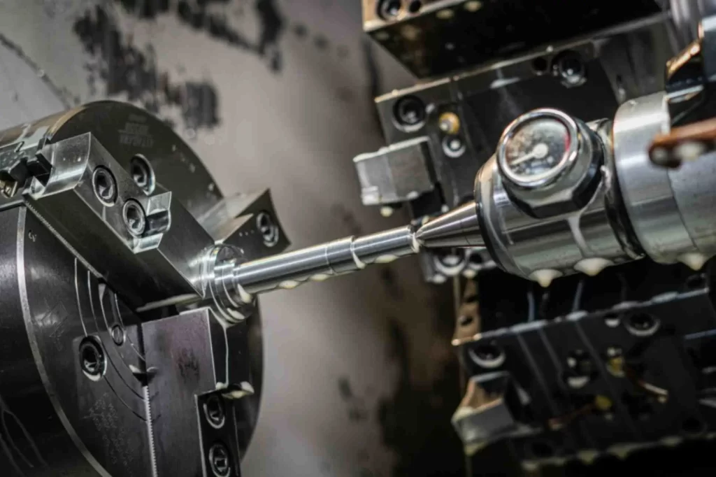 A metallic rod with varying cross-sections held by a CNC machine being machined to reduce the diameter