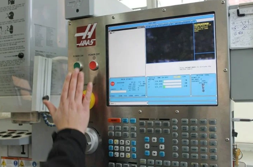 A closeup of a CNC machine control panel showing a program on the screen and a person about to press the green button