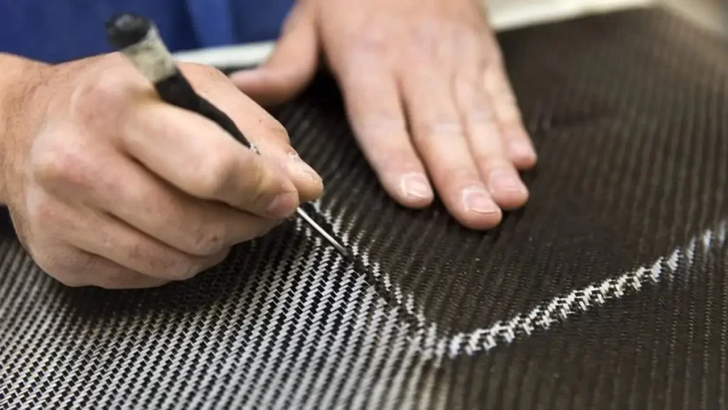 A close-up of a person's hands using a tool to draw on a sheet of carbon fiber material.