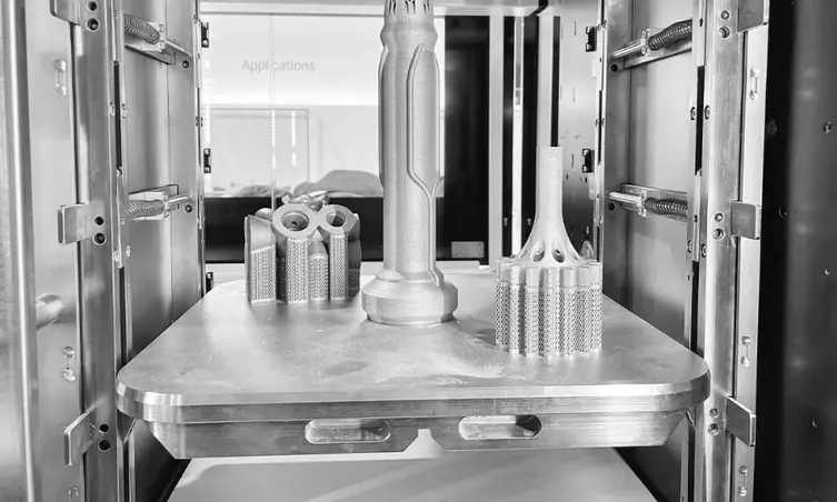 Interior view of industrial 3D printer build chamber showing multiple metal prototype parts on heated build platform