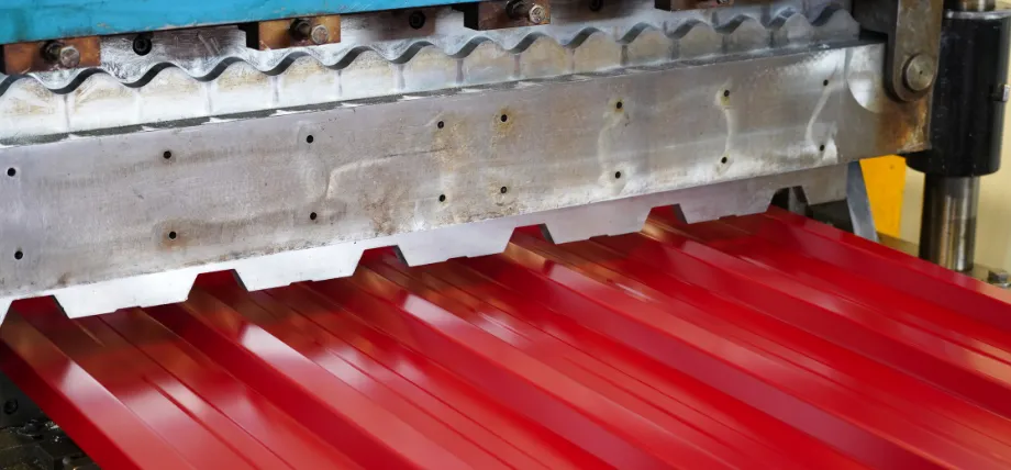 Close-up of a roll-forming machine shaping glossy red corrugated sheet metal as it exits the forming rollers.