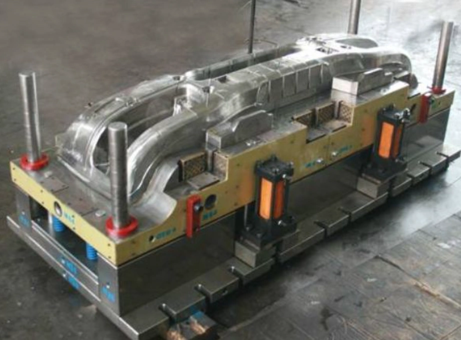 Large-scale injection mold tooling for an automotive plastic bumper, showing cavity, side-action cylinders, and guide pillars on a workshop floor