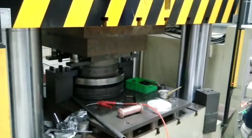 Industrial cold forging process showing a press machine with yellow and black safety striping and metal workpieces on the platform