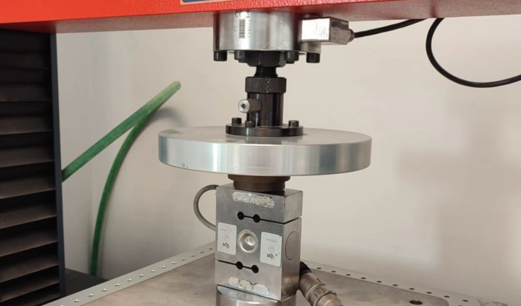 A CNC machined circular metal disc between two parts of a compressive test machine