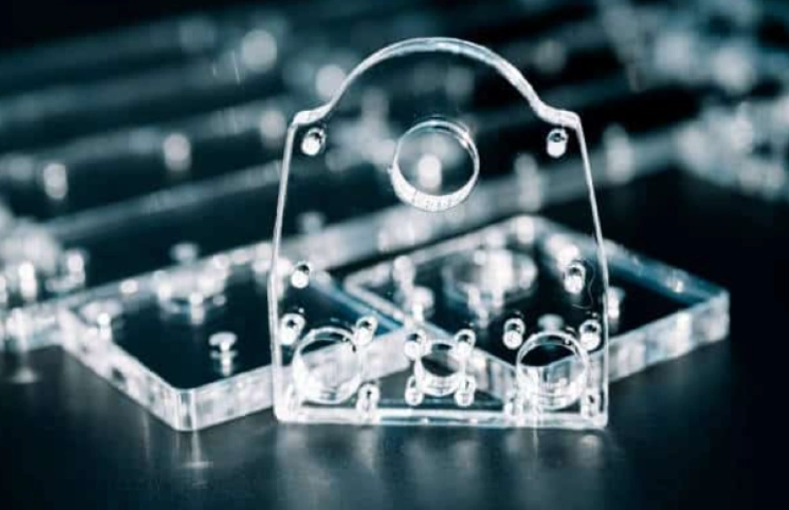 An image of CNC machined transparent parts with varying shapes and holes on a dark reflective surface