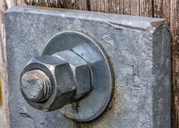 A closeup image of two nuts and a bolt end securing a metal plate on a piece of wood with a flat washer shown